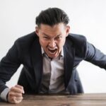 Interdepartmental Disputes - Expressive angry businessman in formal suit looking at camera and screaming with madness while hitting desk with fist